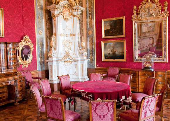 Blick in einen der prunkvoll eingerichteten Kaiserzimmer mit rotem Wandbehang, runden Tisch mit Stühlen.