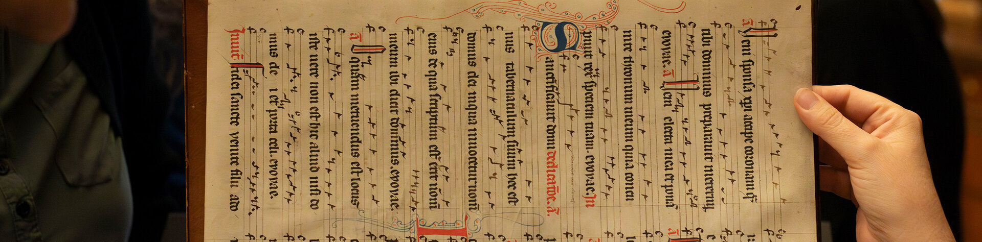 Person hält eine alte Handschrift hoch, im Hintergrund schauen mehrere Menschen zu.