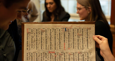 Person hält eine alte Handschrift hoch, im Hintergrund schauen mehrere Menschen zu.