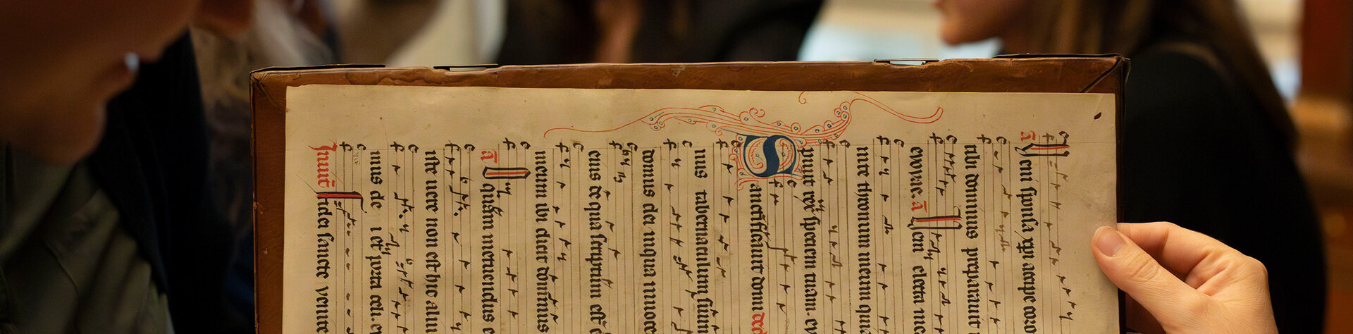 Person hält eine alte Handschrift hoch, im Hintergrund schauen mehrere Menschen zu.