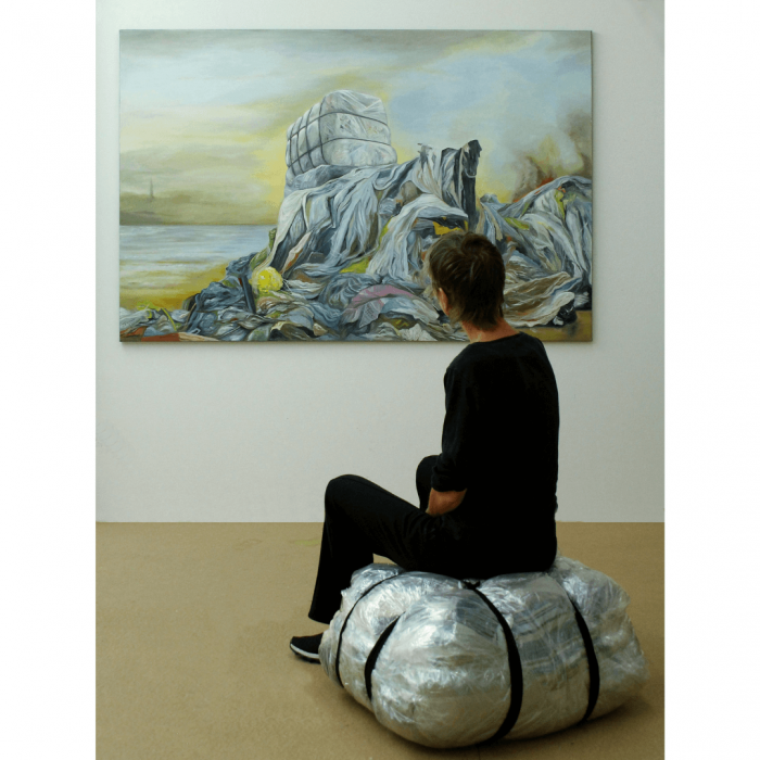 Gemälde zeigt Textilmüll, davor sitzt eine Frau auf einem in Plastik gewickelten Altkleiderballen